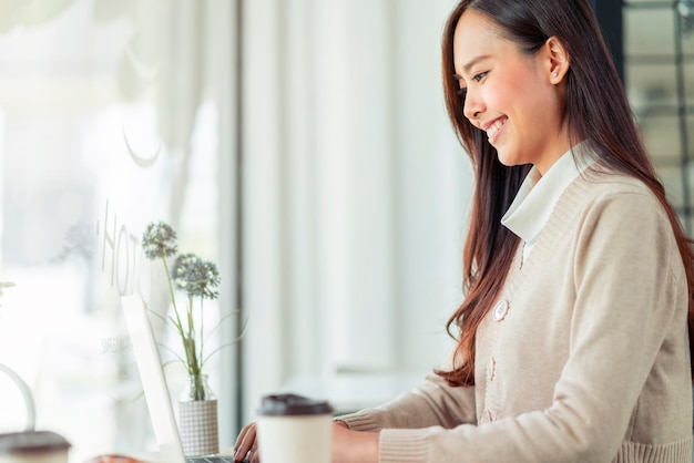 La bella mattina allegra di sorriso femminile asiatico attraente di freschezza che lavora con il lavoro del caffè del computer portatile da casa con la felicità gode della giovane donna asiatica di vista del giardino con la priorità bassa del giardino della luce della finestra