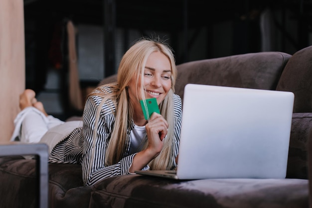 La bella modella bionda sdraiata sul divano con il computer portatile a casa tiene la carta di credito effettua l'acquisto online guarda lo schermo soddisfatto dalla vendita di sconto Venerdì nero La donna di successo acquista via internet