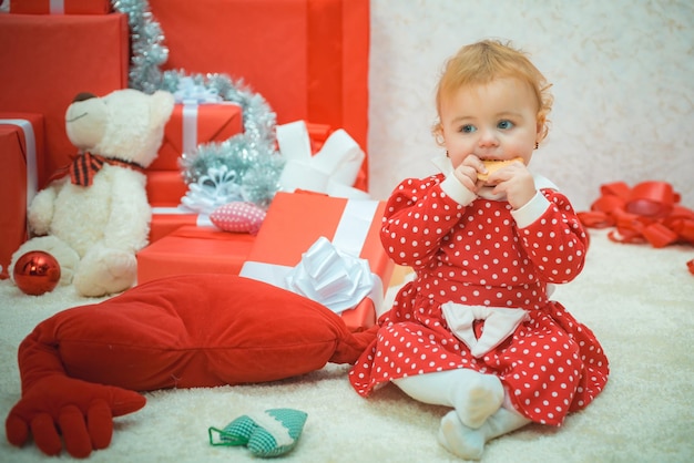 La bella neonata in vestito rosso sta mangiando i biscotti sulla purezza del bambino adorabile del fondo dei regali di Natale