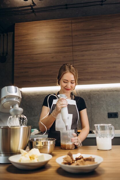 La bella pasticcera vestita con grembiule batte la crema con il mixer nella cucina moderna