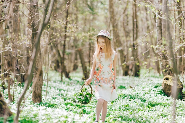 La bella piccola ragazza bionda cammina nella foresta di primavera. Ritratto di una bella bambina con un cappello in testa e un cesto con bucaneve