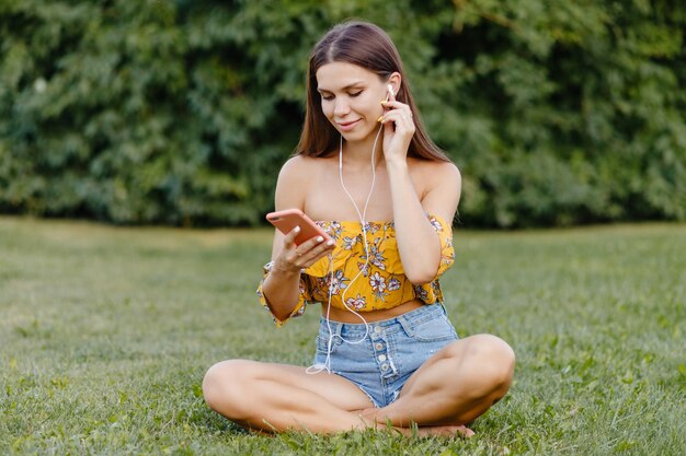 La bella ragazza ascolta la musica al telefono mentre si siede sull'erba nel parco