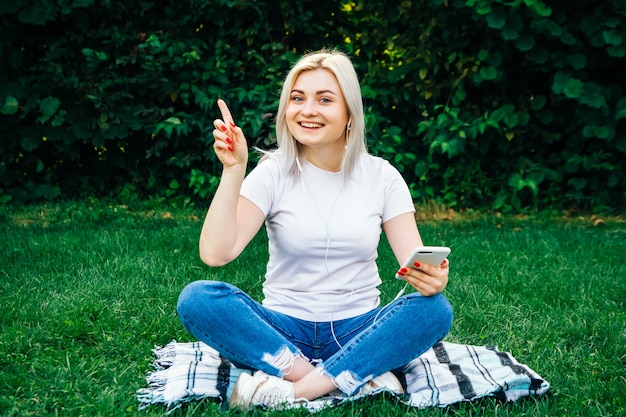 La bella ragazza bionda in cuffie e smartphone in mani si siede sull'erba verde