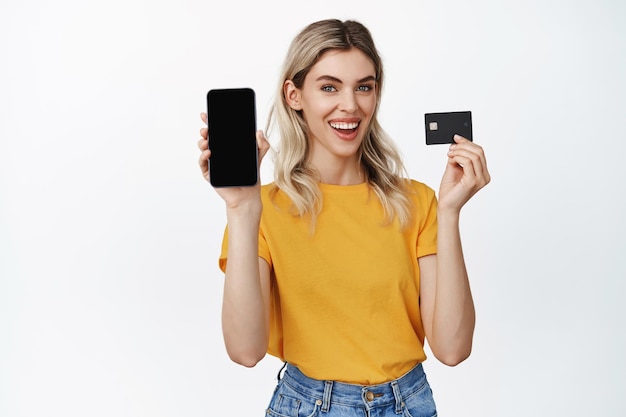 La bella ragazza bionda mostra lo schermo del telefono cellulare con la carta di credito che consiglia l'interfaccia dell'app per smartphone dell'applicazione in piedi in maglietta gialla su sfondo bianco