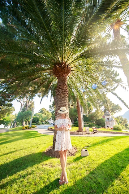 La bella ragazza brillante con un vestito leggero e un cappello in piedi sotto una palma di monaco in un clima soleggiato