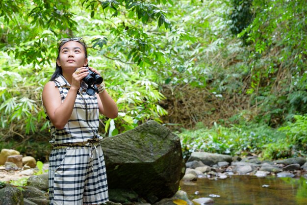 La bella ragazza che fa un'escursione sta usando il binocolo per cercare gli uccelli nella foresta tropicale vicino alle cascate in Thailandia