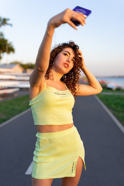 La bella ragazza con i riccioli in una gonna verde e un top si fa un selfie sotto i raggi del sole della sera