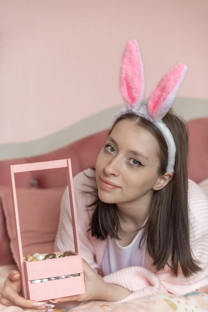 La bella ragazza con le orecchie del coniglietto sulla sua testa si trova sul letto a casa con un cestino delle uova di Pasqua