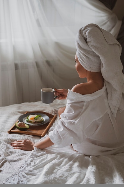 La bella ragazza con un asciugamano bianco in testa si siede a letto la mattina e beve caffè