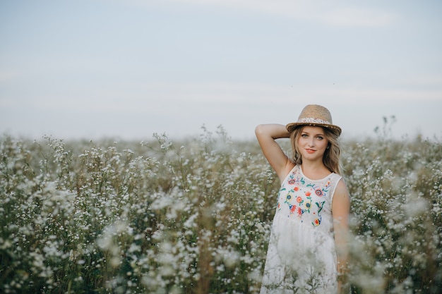 La bella ragazza con un cappello in mano cammina in un campo con fiori di campo e sorride sinceramente