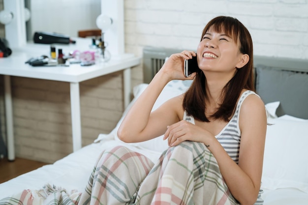 la bella ragazza coreana a testa in su e mettendo una mano sul ginocchio sta ridendo per una battuta della sua amica al telefono. la giovane femmina asiatica che guarda verso l'alto sta chattando felicemente sul cellulare seduto sul letto