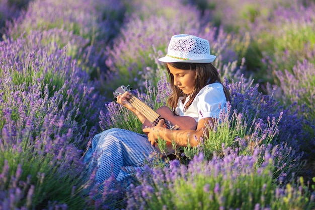 La bella ragazza di campagna in un cappello suona una piccola chitarra mentre è seduto in un campo di lavanda