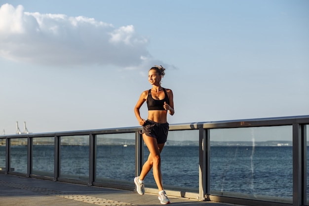 La bella ragazza di forma fisica in pantaloncini corti e sport top corre sul lungomare in riva al mare durante il tramonto