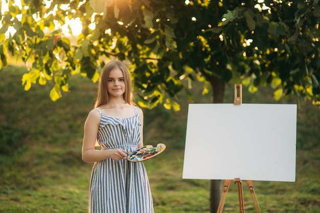 La bella ragazza disegna un'immagine nel parco usando una tavolozza con vernici e una spatola. Cavalletto e tela con una foto.