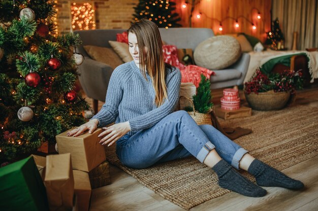 La bella ragazza felice allegra stabilisce i regali di Natale sul pavimento vicino all'albero di nuovo anno a casa