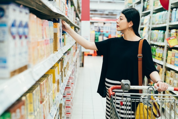 la bella ragazza genitore giapponese asiatica futura prende da bere dagli scaffali del dipartimento delle bevande del supermercato. signora incinta che fa shopping nell'ipermercato. vista laterale maternità spingendo il carrello nel negozio di alimentari