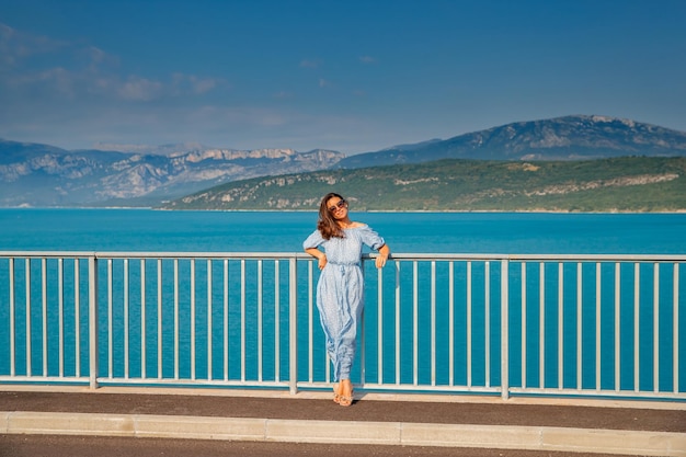 La bella ragazza in abito blu e occhiali da sole posa sul ponte i lunghi capelli castani felice a