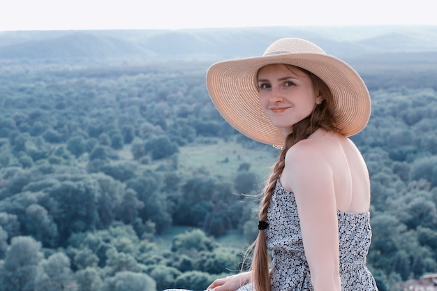 La bella ragazza in cappello e vestito si siede su una foresta dell'estate. Tonificante