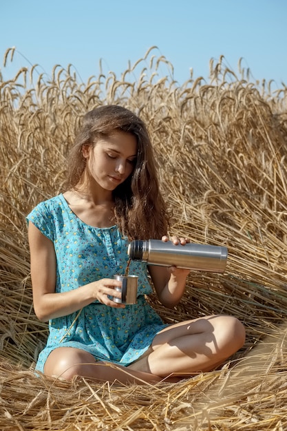 La bella ragazza in prendisole blu si siede sul campo di frumento e beve il tè o il caffè dalla boccetta del thermos