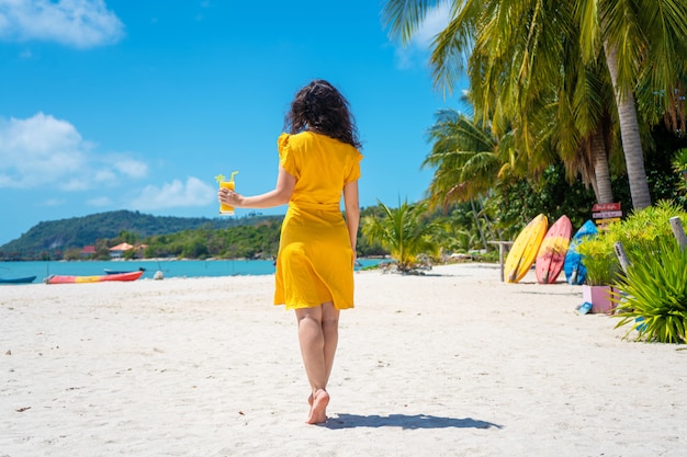 La bella ragazza in un vestito giallo beve il mango fresco sulla spiaggia di un'isola di paradiso. Vacanza perfetta.