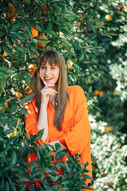 La bella ragazza in vestito arancione sta posando alla macchina fotografica mettendo la mano sotto il mento nel giardino degli aranci