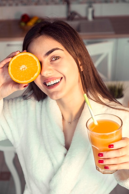 La bella ragazza indossa l'accappatoio bianco con un bicchiere di succo d'arancia e la vista del tramonto mattutino della cucina della frutta