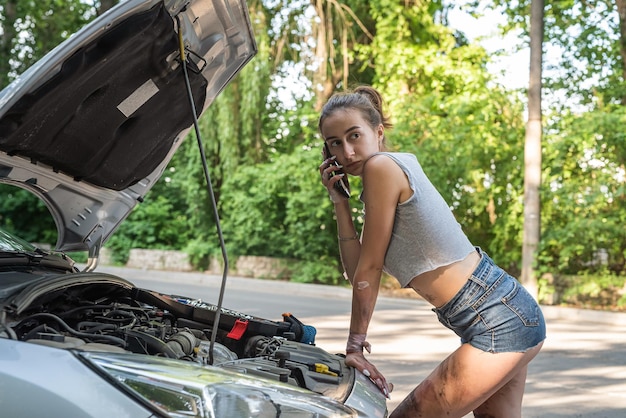 La bella ragazza indossa pantaloncini corti e magliette che esaminano il vano motore delle vacanze estive in auto in panne