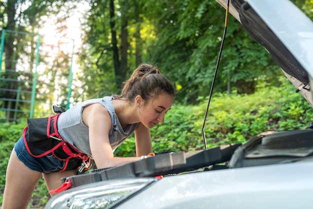 La bella ragazza indossa pantaloncini corti e magliette che esaminano il vano motore delle vacanze estive in auto in panne