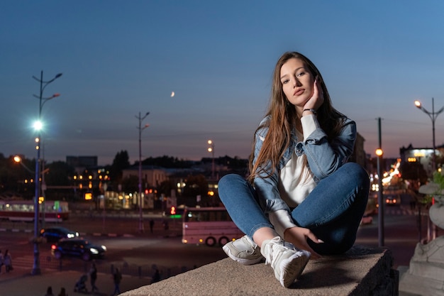 La bella ragazza si siede sullo sfondo della città di sera. Elegante studentessa sola seduta sul gradino alto.