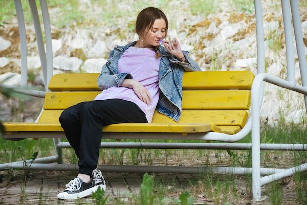 La bella ragazza sognante si siede su una panchina gialla nel parco nella stagione calda, si gode una passeggiata