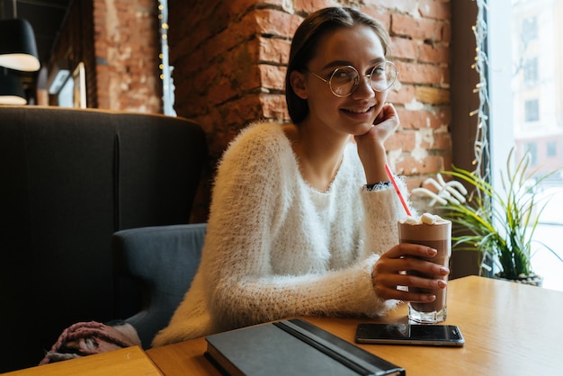 La bella ragazza sorridente freelance con una giacca bianca e occhiali si siede in un caffè a un tavolo e beve un latte profumato