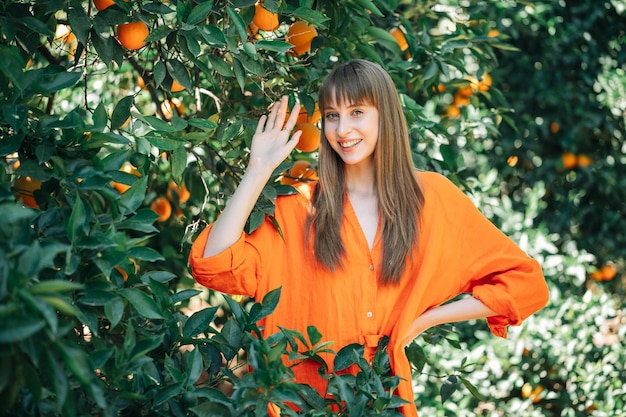 La bella ragazza sorridente in vestito arancione sta mostrando ciao gesto e mettendo l'altra mano sulla vita nel giardino d'arancio