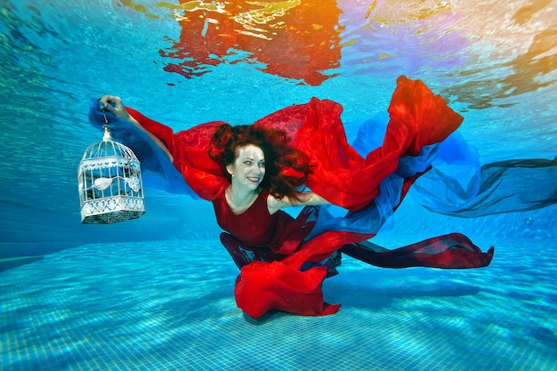 La bella ragazza sott'acqua nuota e gioca con il tessuto rosso e blu sul fondo della piscina