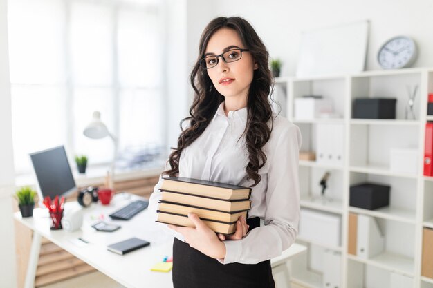 La bella ragazza sta nell'ufficio e tiene una pila di libri in sue mani.