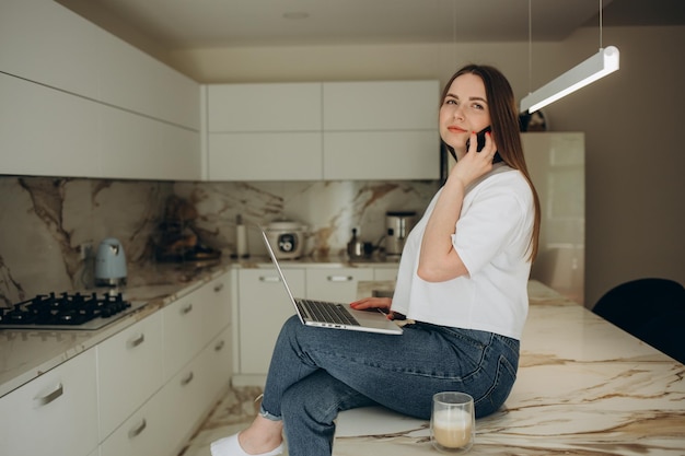 La bella ragazza sta parlando al telefono cellulare che tiene una tazza che distoglie lo sguardo e sorride mentre è seduto sul tavolo in cucina