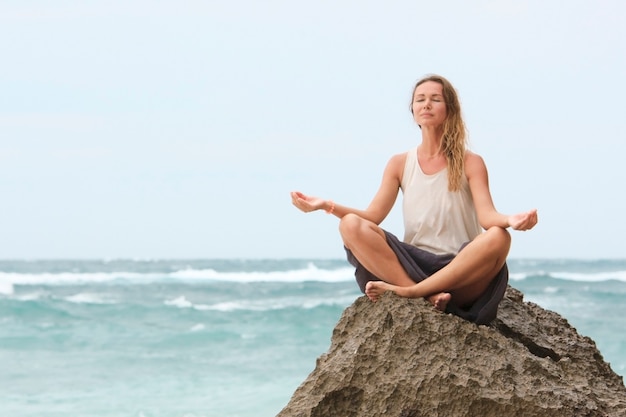 La bella ragazza vestita di bianco si siede in riva al mare sulla roccia e medita nella posa della donna yoga