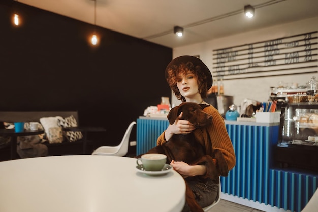 La bella signora in abiti vintage si siede a tavola in un caffè amico degli animali con un cane in braccio