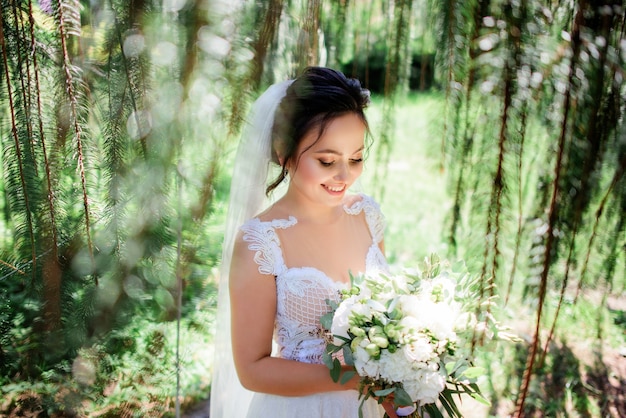 La bella sposa in un vestito tenero posa con il mazzo di nozze nella foresta verde