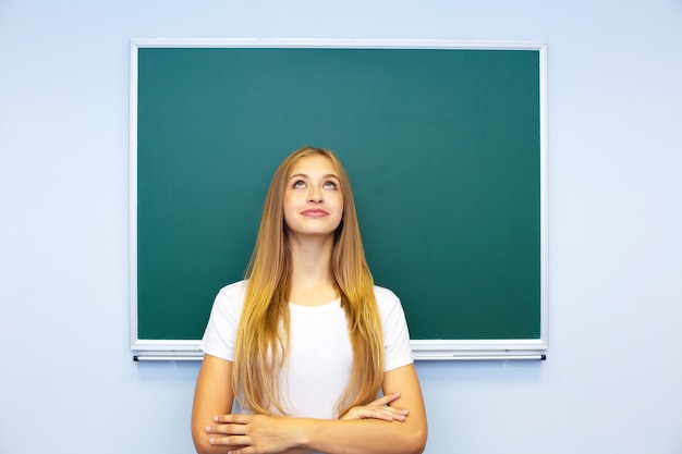 La bella studentessa felice sta vicino alla lavagna e guarda pensieroso verso l&#39;alto.