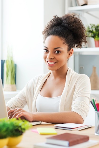 La bellezza al lavoro. Bella giovane donna africana che lavora al computer portatile e sorride mentre è seduta al suo posto di lavoro in ufficio