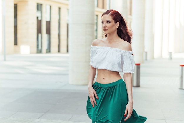 La bellissima modella dai capelli rossi con una camicetta e una gonna verde sta camminando vicino a un edificio alla moda.