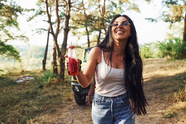 La bionda con l'alcol in mano fa una passeggiata nella foresta contro la jeep verde.