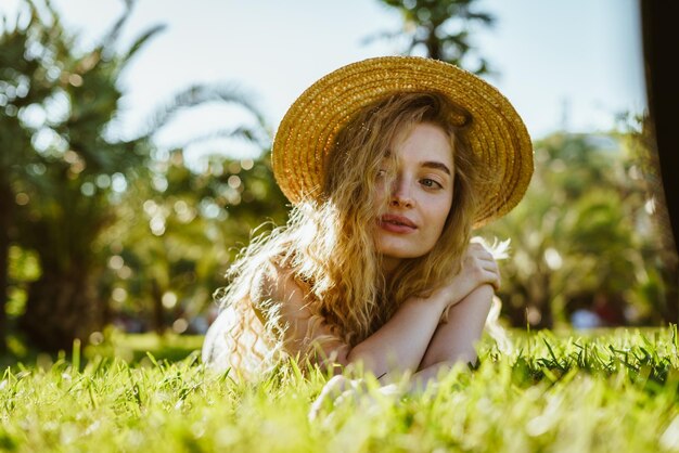 La bionda riccia che sembra una fata cowgirl con un cappello di paglia giace sull'erba nel parco