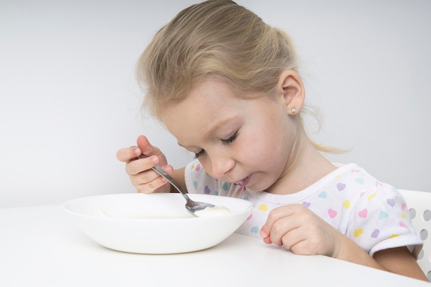 La bionda triste della ragazza del bambino mangia il porridge ad un tavolo bianco