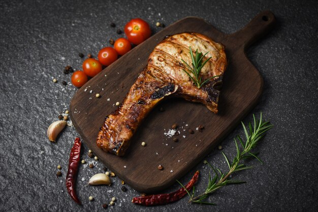 La bistecca di braciole di maiale con l'erba e le spezie serve con la verdura sul bordo di legno - carne arrostita di braciola di maiale su superficie nera