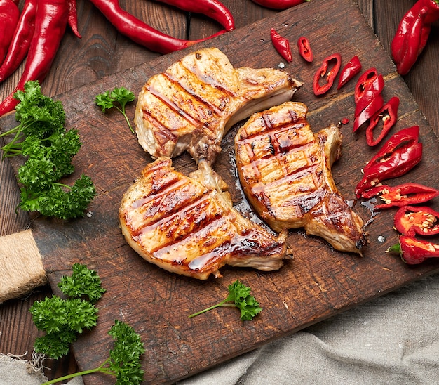 La bistecca fritta carne di maiale sulla costola si trova su un bordo di legno marrone d'annata, accanto ai peperoncini rossi freschi, vista superiore