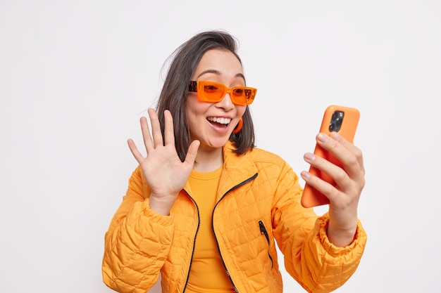 La blogger femminile alla moda positiva saluta i follower nei social network indossa la giacca arancione e gli occhiali da sole fanno il gesto di saluto allo smartphone isolato sul muro bianco si collega alla chat degli amici