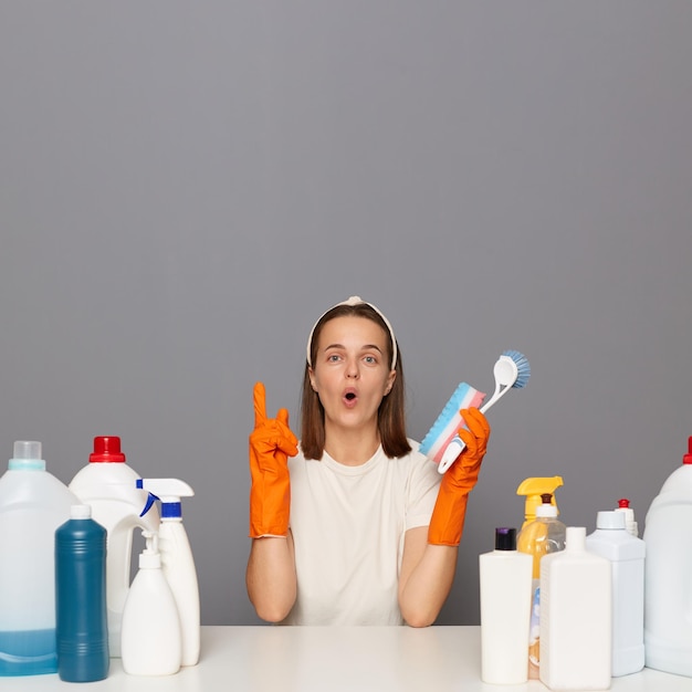 La cameriera eccitata indossa la fascia e la maglietta casual rivolta verso l'alto con il dito indice isolato su sfondo grigio che mostra qualcosa di fantastico sul soffitto utilizza vari detergenti