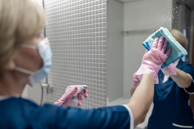 La cameriera in maschera pulisce lo specchio nel bagno dell'hotel spruzzando detergente sulla superficie