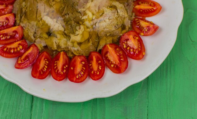 La carne in gelatina in un piatto rotondo decorato con pomodori su una superficie di legno di colore verde chiaro